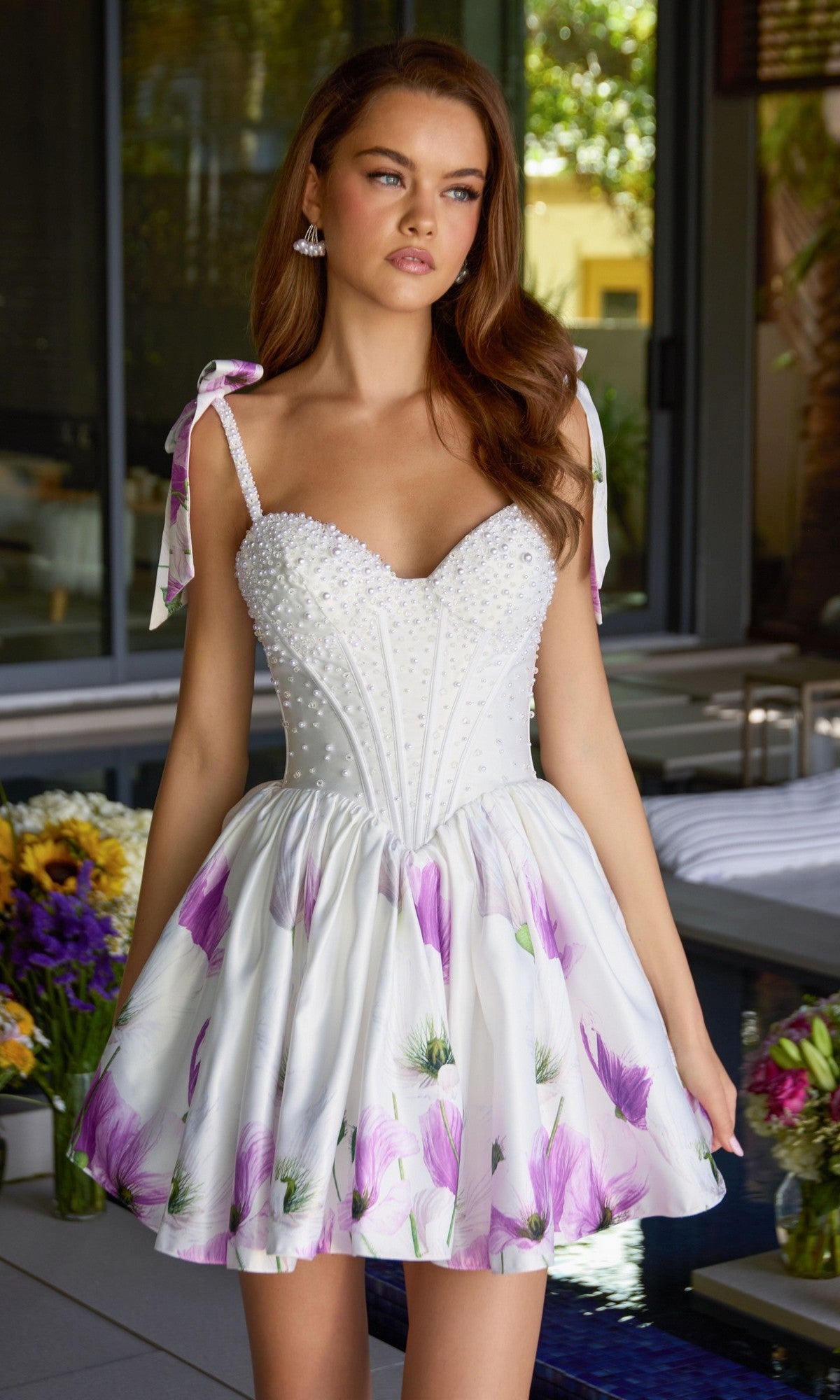 A woman in the Ava Presley 42432 short cocktail dress stands indoors near a pool and flower arrangements. The designer dress features a floral satin print and a pearl-embellished corset.
